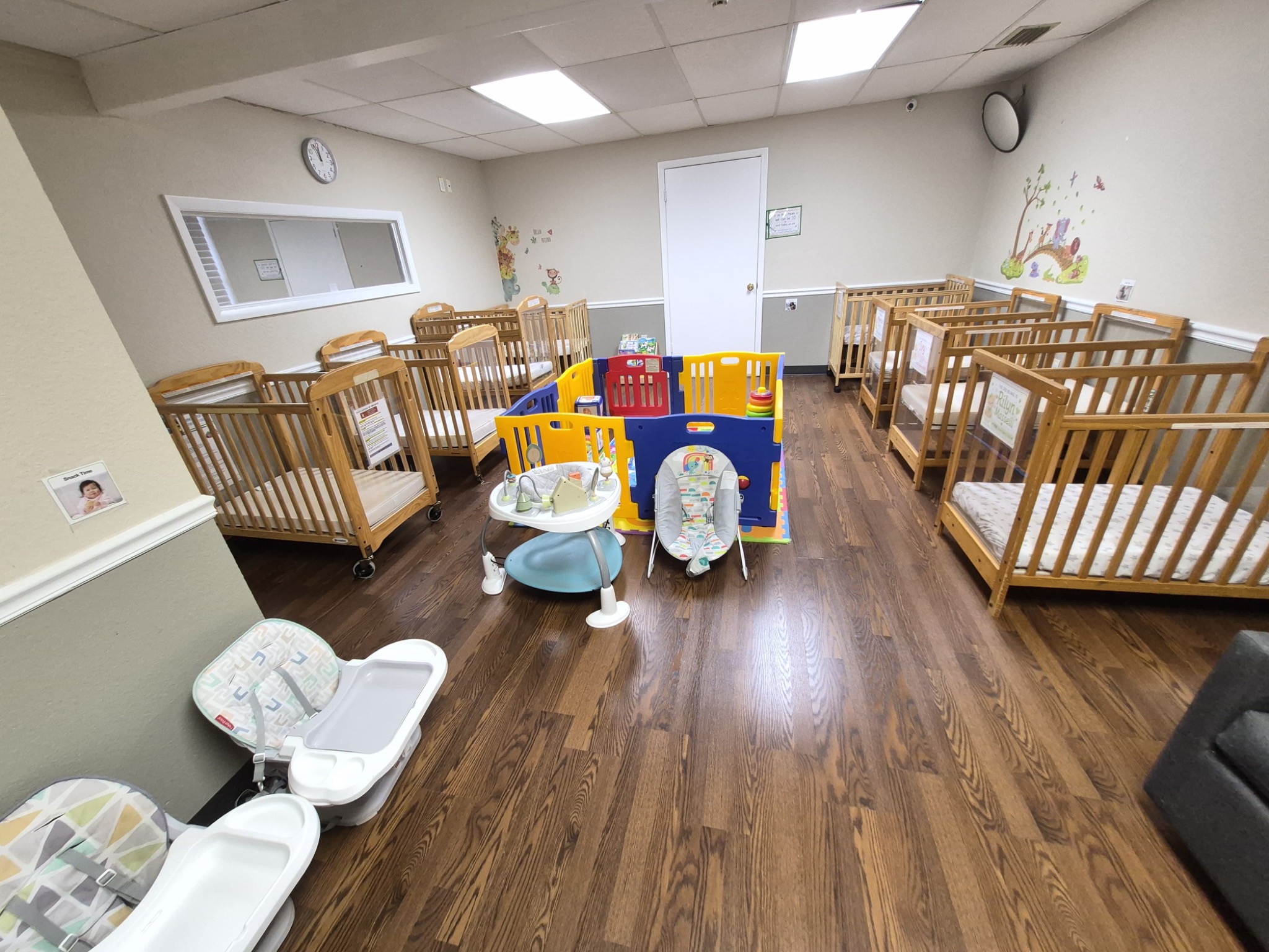 Infant room with wooden cribs, wood flooring, baby seats and colorful play equipment